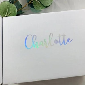 Puede incluir: Caja de regalo blanca con un dise&ntilde;o hologr&aacute;fico iridiscente. La caja tiene el nombre "Charlotte" impreso en la parte delantera en una fuente cursiva.