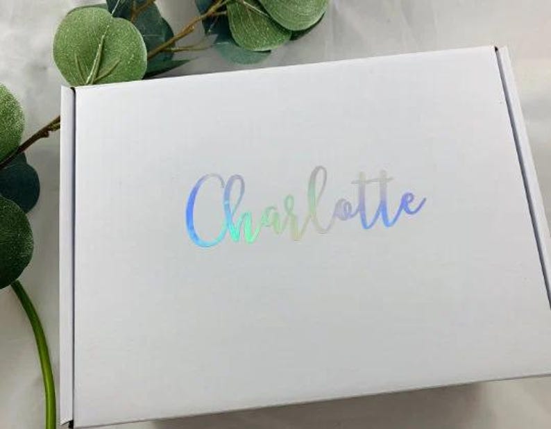 Puede incluir: Caja de regalo blanca con un dise&ntilde;o hologr&aacute;fico iridiscente. La caja tiene el nombre "Charlotte" impreso en la parte delantera en una fuente cursiva.