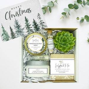 May include: A white gift box filled with a variety of items, including a small succulent plant, a candle, a bar of soap, and a small jar of matches. The box is decorated with a white and green watercolor design of pine trees and the words "Merry Christmas".
