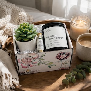 May include: A gift box with a succulent plant in a white pot, a jar of matches, and a black jar candle labeled "Mango + Coconut." The box has floral designs. A lit candle and a cup of coffee are also present.