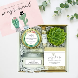May include: A white gift box with a card that says "My wedding would succ without you, be my bridesmaid?"  The box contains a succulent plant, a small jar of mango and coconut scented soy wax candle, a small jar of vanilla scented candle, a small bottle of vanilla lip balm, a small bottle of matches, and a bar of eucalyptus and tea tree soap.