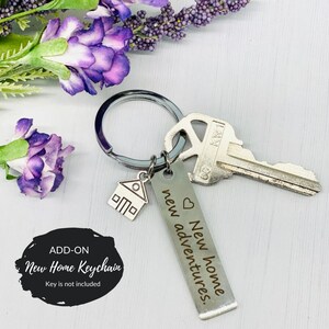 Może przedstawiać: Srebrny brelok do kluczy z zawieszką w kształcie domu, kluczem i srebrną etykietą grawerowaną z napisem "Nowy dom, nowe przygody".