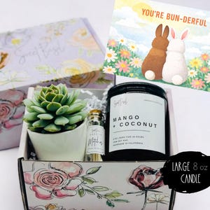May include: Gift box with a succulent, candle, and a small bottle. The candle is labeled "Mango + Coconut" and the box has floral designs. A card with two bunnies and the text "You're Bun-derful" is included.