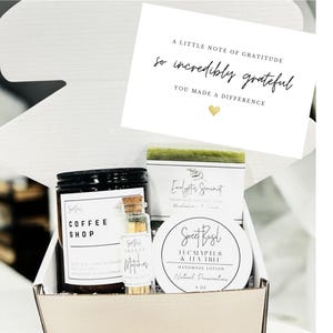 May include: A gift box containing a candle, lotion, soap, and a small bottle of oil. The candle has a black lid and a label that reads "Coffee Shop." A card with the text "A little note of gratitude" is also included.