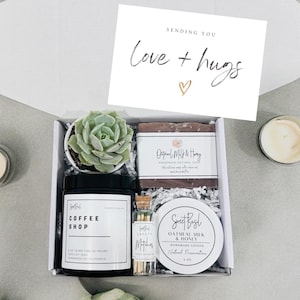 May include: A gift box filled with self-care items, including a candle, soap, matches, lotion, and a small succulent plant. The box is decorated with shredded paper and a card that says "Sending you Love + Hugs".