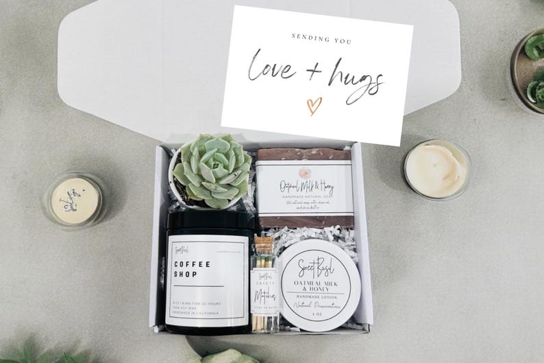 May include: A gift box filled with self-care items, including a candle, soap, matches, lotion, and a small succulent plant. The box is decorated with shredded paper and a card that says "Sending you Love + Hugs".