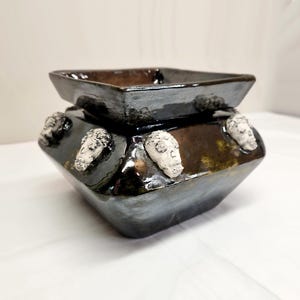 May include: A square ceramic planter with a glossy, dark metallic finish. The exterior features four small, white, sculpted skull-shaped embellishments attached to the sides. The vessel has a wide, open top and a tapered base with a smooth, reflective surface.