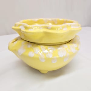 May include: Two-piece ceramic set in a bright yellow color with white accents. The top piece is a shallow bowl with a scalloped edge, and the bottom piece is a larger, rounded bowl with three small feet. The set has a glossy finish.