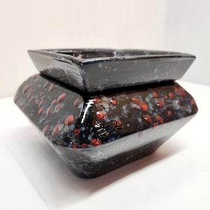 May include: A two-tiered, geometric ceramic container. The dark, glossy surface is speckled with red and white spots. The top section is a shallow tray, while the base is a wider, angular form. The container is likely used for decorative purposes.