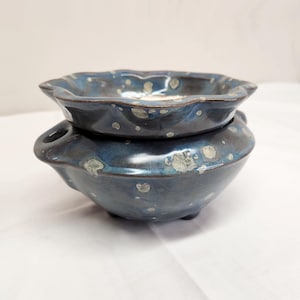 May include: A two-piece ceramic wax warmer featuring a deep blue glaze with mottled grey and white speckles. The top section is a scalloped dish, while the base is a rounded bowl with a small circular opening on the side for a light source.