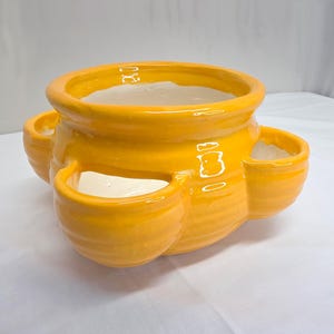 May include: A bright yellow ceramic strawberry planter with a glossy finish. The pot features a large central opening and three smaller side pockets for planting, all with a smooth, rounded rim and a white interior glaze throughout the vessel.