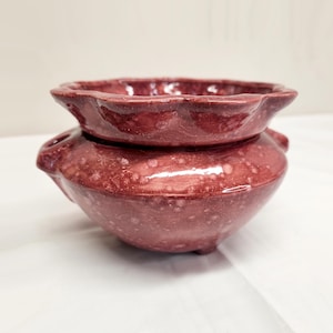 May include: A deep red ceramic bowl with a glossy, speckled finish and a scalloped rim. The bowl features a rounded body with small side handles and sits on a short base, showcasing a smooth, reflective surface with lighter pinkish spots.