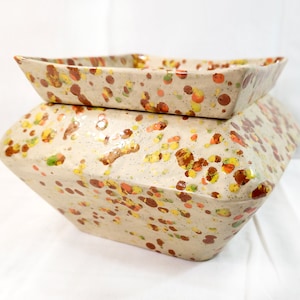 May include: A white ceramic planter with a square top and a geometric base. The planter is decorated with a speckled pattern of brown, orange, and yellow dots.