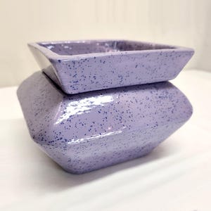 May include: A two-piece ceramic container in a soft lavender hue with a speckled pattern. The top piece is a square shape, and the bottom is a geometric design. The container is likely for home decor or storage.