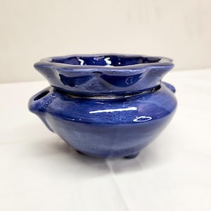 May include: A deep blue ceramic oil warmer with a glossy finish. The piece features a wide, scalloped-edge bowl at the top for wax or oil, a rounded base with a small side opening for a tea light, and three small feet for stability.
