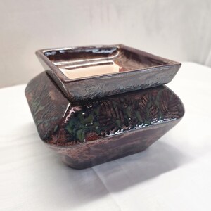 May include: A square ceramic wax warmer featuring a dark brown, metallic-toned glaze with a textured, leaf-like pattern embossed on the sides. The top section includes a removable square dish with a smooth, light-colored interior surface.