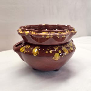 May include: A dark brown ceramic planter with a glossy finish and a scalloped rim. The surface features irregular, bright yellow drip-like spots scattered across the exterior and the upper edge. It has a rounded body and small feet.
