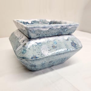 May include: A square ceramic wax warmer featuring a light blue and white speckled glaze with a glossy finish. The piece consists of a base with a tapered shape and a removable square dish on top, both decorated with a consistent mottled pattern.