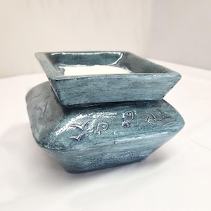 May include: A two-piece teal ceramic container with a square top and a larger, decorative base. The base features embossed bird and wave designs. The top section holds a white substance. The container has a textured, aged appearance.