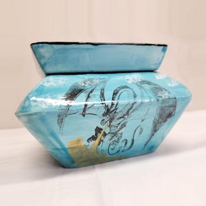 May include: A light blue ceramic jar with a lid, featuring a black ink illustration of a winged dragon breathing golden fire. The vessel has a wide, angular body and a smooth, glossy finish, with white cloud-like accents painted across the surface.