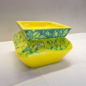 May include: A square ceramic bowl with a bright yellow glaze and a contrasting teal blue leaf pattern imprinted on the exterior and the inner rim. The vessel features a wide, flat opening and a tapered base, showcasing a glossy, smooth finish.