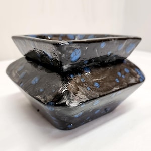 May include: A handmade, square ceramic bowl with a two-tiered design. The bowl is glazed in a glossy black finish with speckled blue accents. The bowl's shape is geometric and modern, suitable for home decor or serving.
