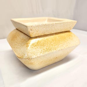 May include: A two-piece, beige ceramic container with a square lid. The base is a larger, geometric shape with rounded edges. The surface has a textured, speckled appearance. The container is likely for storage or decorative purposes.