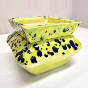 May include: Two square ceramic bowls stacked on top of each other. The bowls are a bright lime green color with dark blue speckled patterns. The bowls have a glossy finish and are empty.