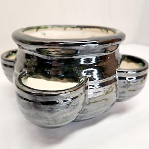 May include: A black and silver ceramic planter with three side pockets. The planter has a glossy finish and a cream-colored interior. The rim of the planter has a green accent.