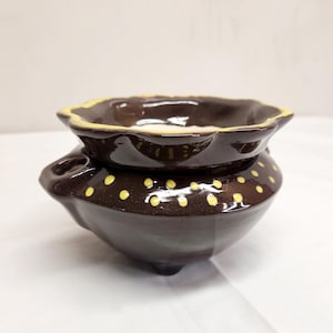 May include: A dark brown ceramic wax warmer with a glossy finish, featuring a scalloped top rim with a yellow edge and a body decorated with small yellow polka dots. The piece has a small side handle and rests on three short, rounded feet.