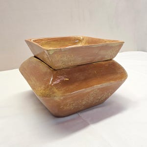 May include: A stack of two square ceramic bowls with a glossy, variegated brown and tan glaze. The larger bottom bowl features a wide, angled rim and a subtle embossed floral scroll pattern on the side, while the smaller top bowl is nested.