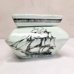 May include: A light green ceramic lidded container with a glossy finish. The lid features a black rope-patterned border. The base displays a detailed black ink illustration of a multi-masted sailing ship on the water, centered on the front.