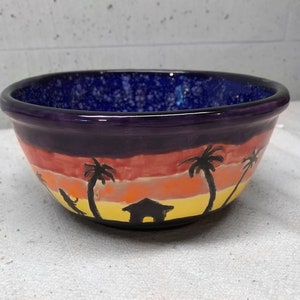 May include: A purple ceramic bowl with a sunset scene painted on the outside. The scene includes palm trees, a small house, and a yellow, orange, and red sunset.