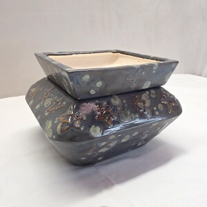 May include: A square ceramic planter with a dark gray glaze featuring light gray mottled spots and embossed star shapes. The planter has a wide, angled base and a separate, smaller square top section with a smooth, light cream-colored interior.