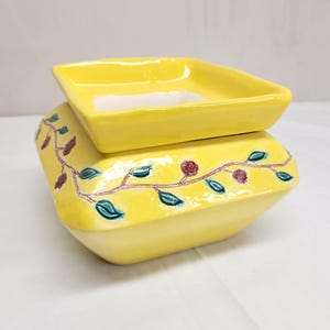 May include: A bright yellow ceramic wax warmer with a square top dish and a wider square base. The base features a hand-painted vine motif with dark green leaves and small reddish-brown berries wrapping around the sides of the glossy yellow surface.