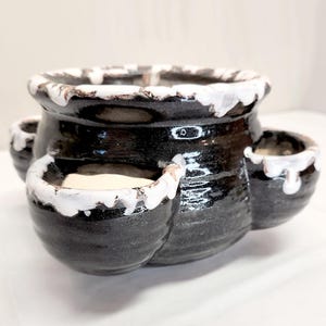 May include: A black ceramic planter with three attached side pots and a wide top opening. The planter has a textured surface with white paint dripping over the edges. The overall design is a unique and decorative piece for plants.