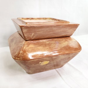 May include: Two-piece ceramic container set. The bottom piece is a large, geometric shape with a wide base and angled sides. The top piece is a square shape with a flat top. Both pieces are painted in a brown and tan color scheme.