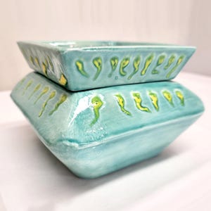 May include: Two-piece ceramic set in a turquoise glaze. The top piece is a rectangular dish, and the bottom piece is a square-based container. Both pieces are decorated with yellow and green accents, creating a unique, handcrafted look.