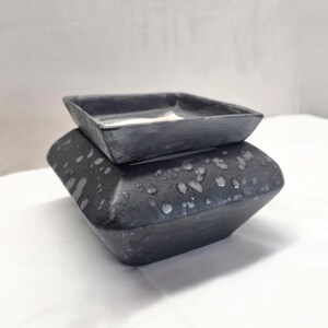 May include: A square ceramic wax warmer featuring a matte black finish with light gray speckled accents. The base has a wide, tapered shape, and the top section is a removable square dish with a smooth interior surface for holding wax melts.