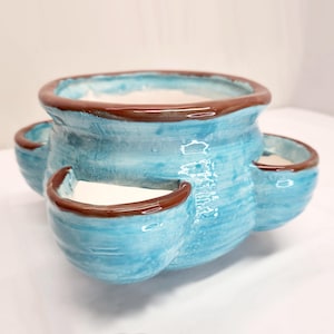 May include: A ceramic planter with a light blue exterior and brown trim. The planter has a round base with three attached side pockets. The interior is white. The planter is designed for growing multiple plants.