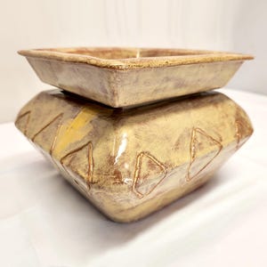 May include: A two-piece ceramic decorative set. The base is a geometric shape with diamond-shaped cutouts and yellow accents. The top is a square dish. The set has a rustic, earthy color palette of tan, brown, and yellow.