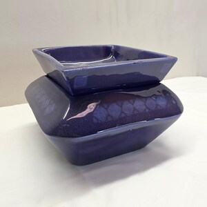 May include: A deep purple ceramic wax warmer with a square, tiered design. The top section is a removable square dish for wax melts, while the base features a wider, angled body with a subtle, darker diamond-patterned glaze finish.