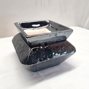 May include: A square ceramic wax warmer featuring a glossy, dark metallic black finish with a textured, dimpled surface on the base. The top section has a smooth, reflective rim and a recessed, off-white interior basin for holding wax melts.
