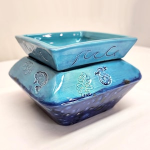 May include: A two-piece ceramic container in shades of blue. The top piece is square with wave-like detailing. The bottom piece has embossed sea life designs, including fish and seaweed. The container has a glossy finish.