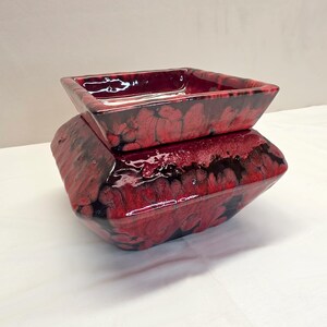 May include: A square ceramic bowl with a glossy finish featuring a marbled pattern of deep red and black. The bowl has a wide, flared rim and a tapered base, showcasing a fluid, organic glaze effect that blends the two colors across the surface.