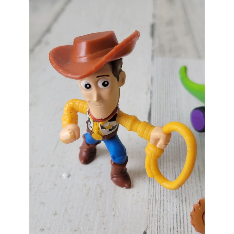 May include: A Woody action figure from the Toy Story franchise. The figure is wearing a brown cowboy hat, yellow shirt, blue pants, and brown boots. He is holding a yellow lasso. The figure has a sheriff's badge on his shirt.