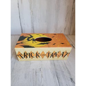 May include: A Halloween-themed tissue box cover with a witch flying on a broomstick in front of a full moon. The cover has a yellow and orange background with bats and stars. The front of the box reads "Trick or Treat".