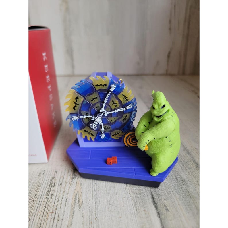May include: A green, stitched, Oogie Boogie character from The Nightmare Before Christmas ornament. He is holding a spinning wheel with a skeleton on it. The wheel is blue and yellow with bats and a spider web design.