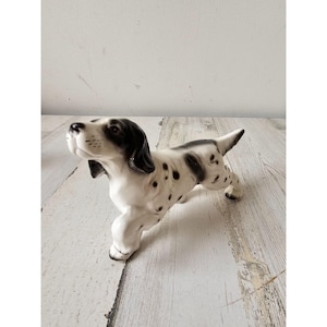 Puede incluir: Una figurita de cerámica de un perro con manchas blancas y negras. El perro está de pie sobre sus cuatro patas con la cabeza girada hacia un lado. La figurita está hecha de cerámica blanca con manchas negras.