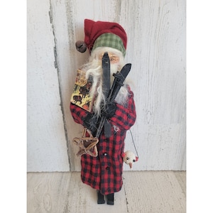 May include: A handmade Santa Claus doll wearing a red and black plaid coat, a green and red hat, and holding a pair of wooden skis. The doll is holding a small book that says "The Polar Express".